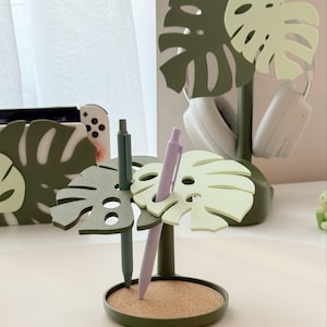 Pu&ograve; includere: Un portapenne a forma di foglia di monstera verde e verde chiaro, con base in sughero. Due penne, una verde e una lavanda, sono inserite. Un motivo a foglia di monstera abbinato &egrave; visibile sullo sfondo, insieme a delle cuffie bianche.