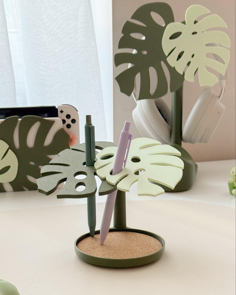 Pu&ograve; includere: Un portapenne a forma di foglia di monstera verde e verde chiaro, con base in sughero. Due penne, una verde e una lavanda, sono inserite. Un motivo a foglia di monstera abbinato &egrave; visibile sullo sfondo, insieme a delle cuffie bianche.