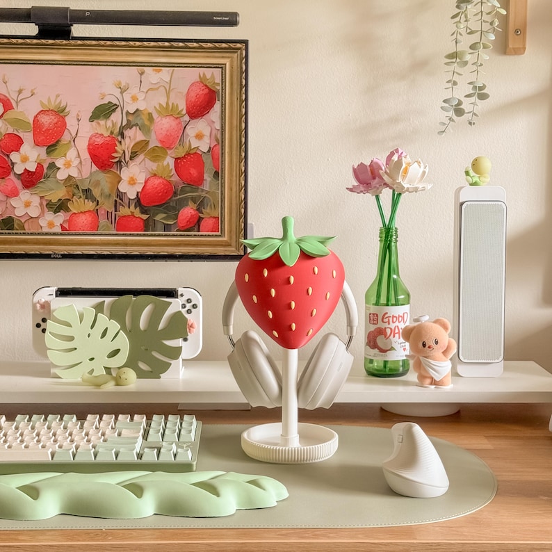 May include: A red strawberry-shaped headphone stand with white headphones. The stand is on a white base. A painting of strawberries hangs on the wall. Other items include a keyboard, mouse, and a green bottle.