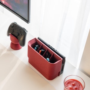May include: A red gaming console organizer with multiple compartments, holding game cartridges and a console. A black controller rests on a matching red stand. A glass of red liquid sits nearby. A black TV is visible in the background.