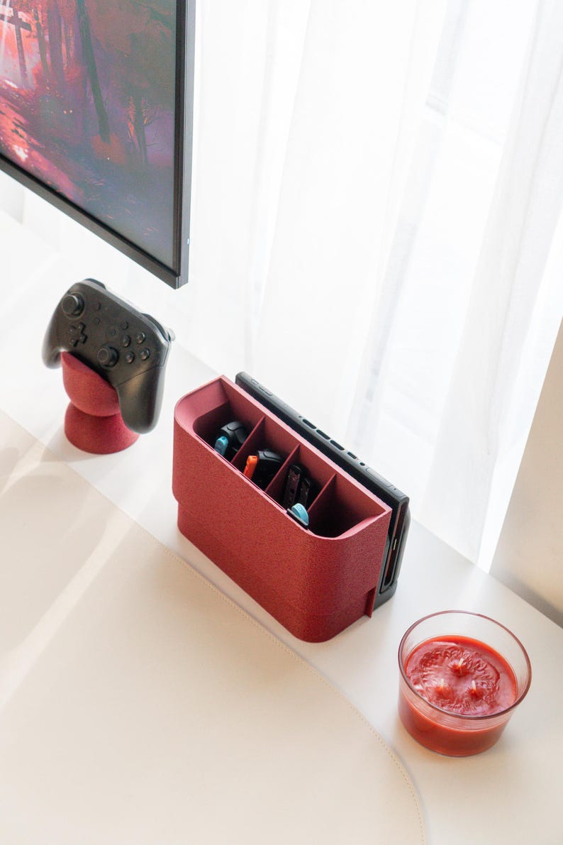 May include: A red gaming console organizer with multiple compartments, holding game cartridges and a console. A black controller rests on a matching red stand. A glass of red liquid sits nearby. A black TV is visible in the background.