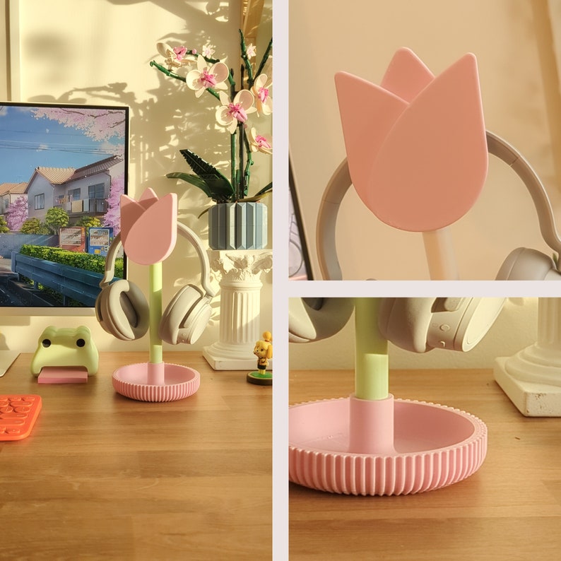 Puede incluir: Un soporte para auriculares en forma de flor rosa con un tallo verde. El soporte tiene una base con una superficie texturizada. Dos auriculares negros est&aacute;n en el soporte.