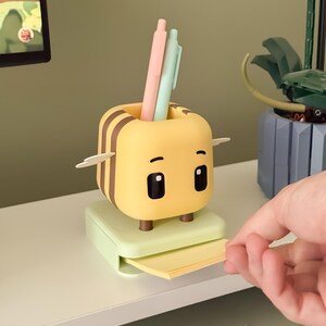 May include: A yellow and brown bee-shaped desk organizer with a pen holder and a small drawer for sticky notes. The organizer has two pens, one pink and one mint green, inside the pen holder. The organizer is sitting on a white shelf.