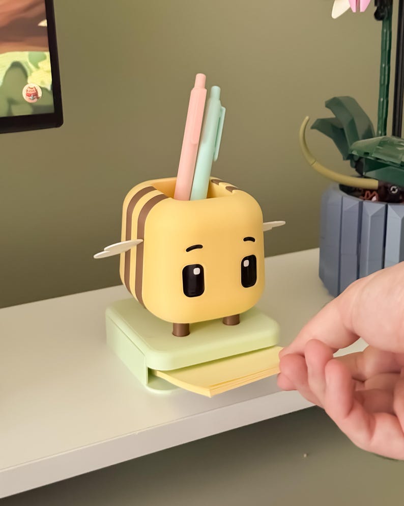 May include: A yellow and brown bee-shaped desk organizer with a pen holder and a small drawer for sticky notes. The organizer has two pens, one pink and one mint green, inside the pen holder. The organizer is sitting on a white shelf.