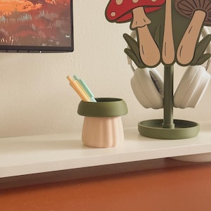 May include: A light pink and green desk organizer shaped like a mushroom, holding pens and pencils. A matching mushroom-themed headphone stand is behind it. The items are on a white shelf against a neutral wall.