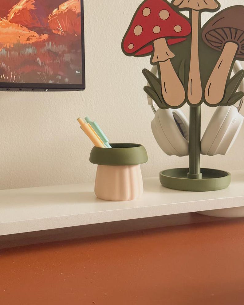 May include: A light pink and green desk organizer shaped like a mushroom, holding pens and pencils. A matching mushroom-themed headphone stand is behind it. The items are on a white shelf against a neutral wall.