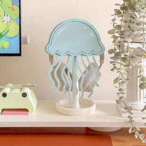 May include: A light blue jellyfish-shaped headphone stand. The stand has a round base and a central support, with the jellyfish design at the top. The headphones are held by the jellyfish's tentacles. The stand is on a white shelf.