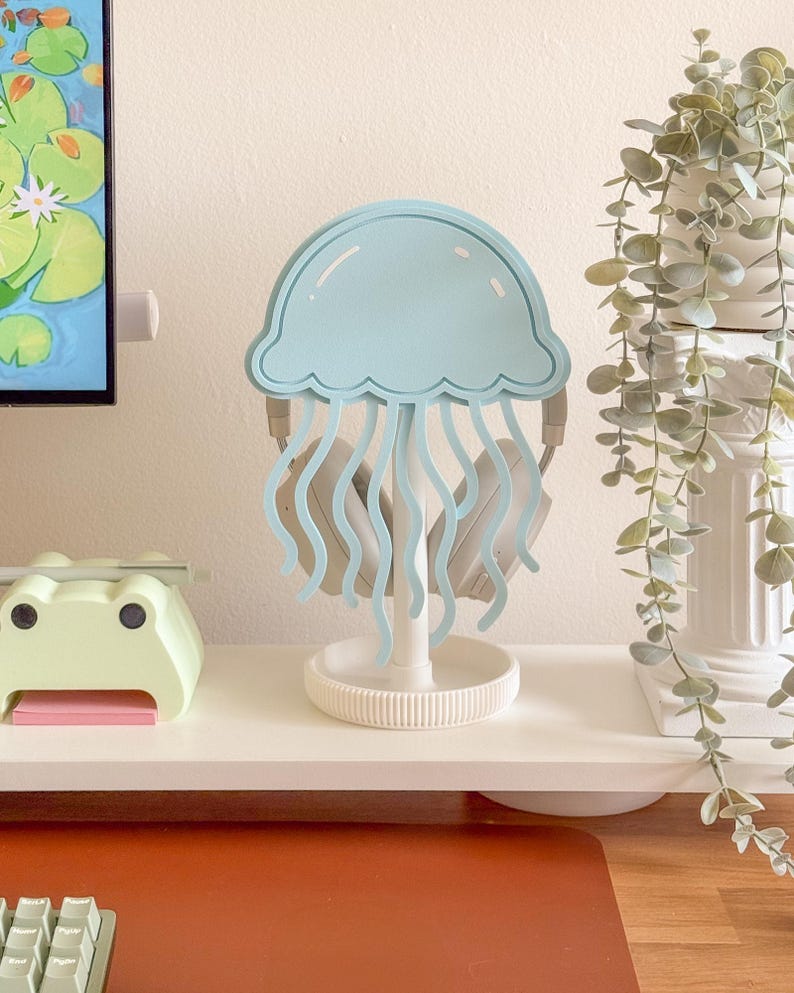 May include: A light blue jellyfish-shaped headphone stand. The stand has a round base and a central support, with the jellyfish design at the top. The headphones are held by the jellyfish's tentacles. The stand is on a white shelf.