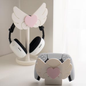 May include: White headphone stand and controller holder, both featuring a wing design with a pink heart accent. The stand holds a pair of white and black headphones. The controller holder is designed for a white game controller.