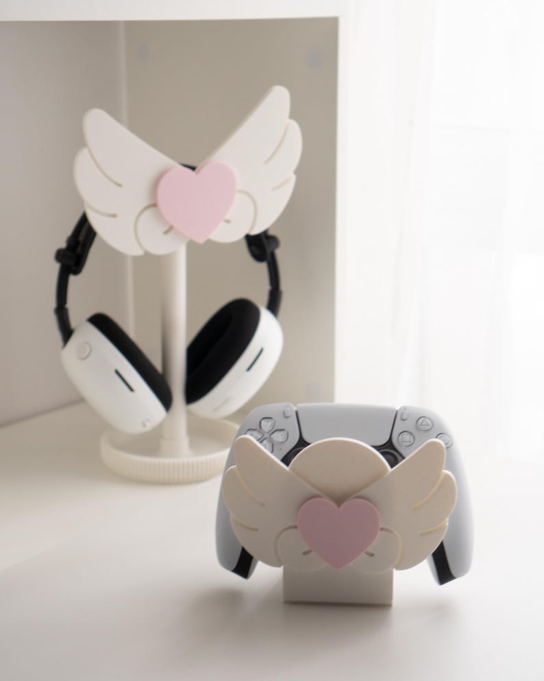 May include: White headphone stand and controller holder, both featuring a wing design with a pink heart accent. The stand holds a pair of white and black headphones. The controller holder is designed for a white game controller.