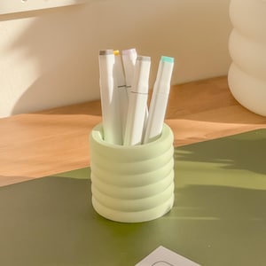 May include: A light green, ribbed pen holder filled with white markers with colored tips. The pen holder sits on a green surface, with a wooden surface in the background. The markers have gray, yellow, purple, and teal tips.