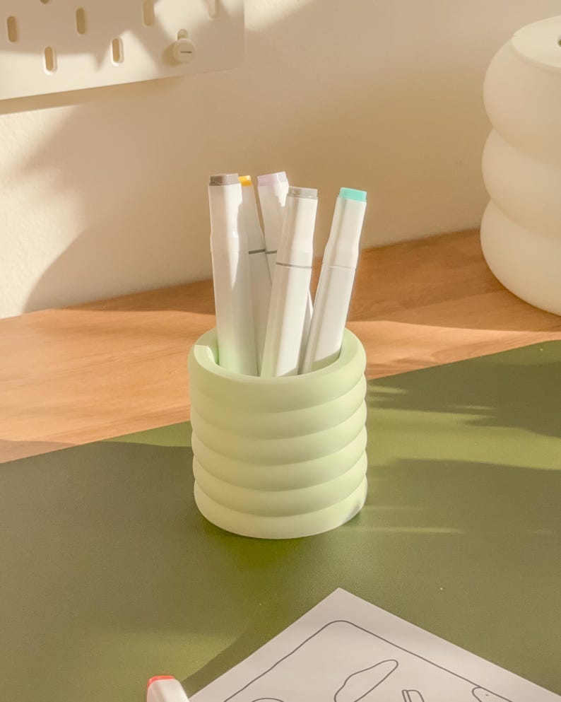 May include: A light green, ribbed pen holder filled with white markers with colored tips. The pen holder sits on a green surface, with a wooden surface in the background. The markers have gray, yellow, purple, and teal tips.