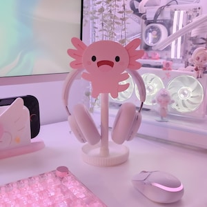 May include: A pink axolotl-shaped headphone stand holding white headphones. The stand is on a white desk with a keyboard and mouse. The background includes a computer and decorative plants.