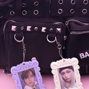 May include: Two decorative photo frame keychains. One is lavender, the other is white. Each frame holds a small photo of a person. The keychains hang in front of a black bag with silver hardware and studded accents.