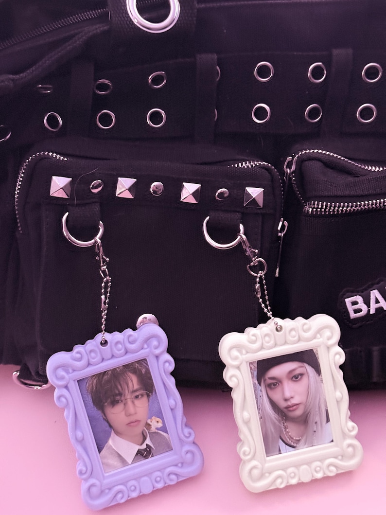May include: Two decorative photo frame keychains. One is lavender, the other is white. Each frame holds a small photo of a person. The keychains hang in front of a black bag with silver hardware and studded accents.