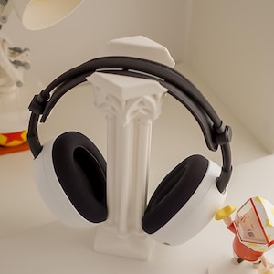 May include: White and black headphones resting on a white decorative column. The headphones have black earcups and a black headband. A small figurine is visible in the background.