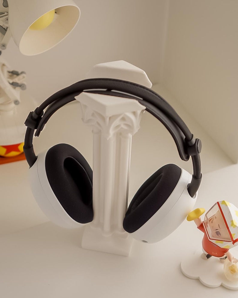May include: White and black headphones resting on a white decorative column. The headphones have black earcups and a black headband. A small figurine is visible in the background.