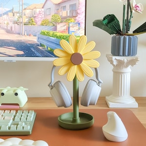 May include: A yellow daisy-shaped headphone stand with two gray headphones hanging from it. The stand is on a brown surface with a green base.