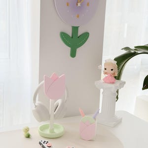 May include: A collection of decorative items, including a tulip-shaped clock, a pink tulip headphone stand, a tulip-shaped pen holder, a small figurine, and two white game controllers. The color palette is soft, with pastel pink, green, and white.