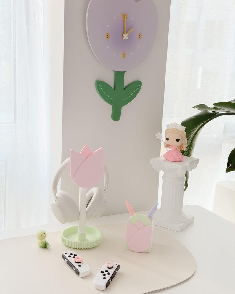 May include: A collection of decorative items, including a tulip-shaped clock, a pink tulip headphone stand, a tulip-shaped pen holder, a small figurine, and two white game controllers. The color palette is soft, with pastel pink, green, and white.