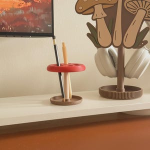 May include: A red and brown desk organizer with a mushroom theme. The organizer holds pencils and pens. A matching mushroom-shaped headphone stand is also visible, along with a television in the background.