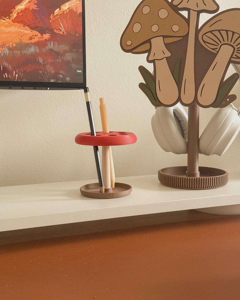 May include: A red and brown desk organizer with a mushroom theme. The organizer holds pencils and pens. A matching mushroom-shaped headphone stand is also visible, along with a television in the background.