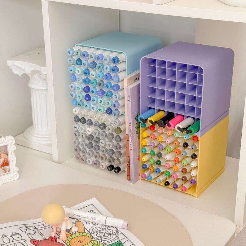 May include: Two rectangular plastic desk organizers with grid compartments. The left unit is light blue and filled with blue and gray markers. The right unit is purple and yellow, containing a variety of colorful markers and pens.