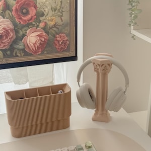 May include: A light pink headphone stand with a decorative column design holds a pair of white headphones. A light brown desk organizer with multiple compartments sits to the left. A floral painting hangs in the background.
