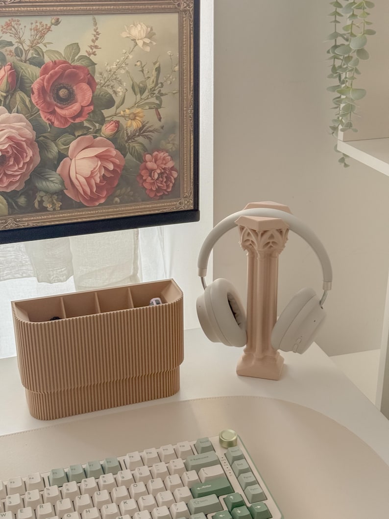 May include: A light pink headphone stand with a decorative column design holds a pair of white headphones. A light brown desk organizer with multiple compartments sits to the left. A floral painting hangs in the background.