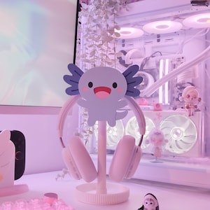 May include: A light pink headset rests on a white stand with a cartoon axolotl design. The stand is on a white desk, with a small penguin figurine and other decorative items in the background. The overall aesthetic is soft and pastel.