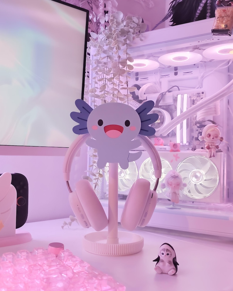 May include: A light pink headset rests on a white stand with a cartoon axolotl design. The stand is on a white desk, with a small penguin figurine and other decorative items in the background. The overall aesthetic is soft and pastel.
