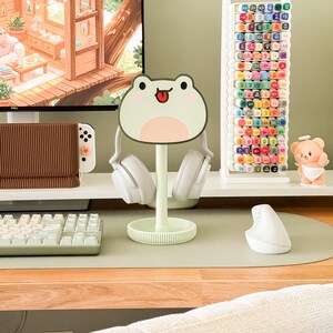 May include: A mint green frog-shaped headphone stand with white headphones. A white marker holder is filled with colorful markers. A small bear figurine sits on a white shelf. A computer monitor displays a cozy cabin scene.