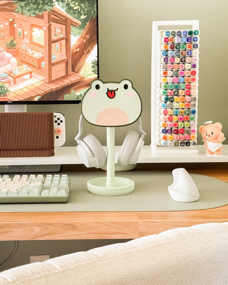 May include: A mint green frog-shaped headphone stand with white headphones. A white marker holder is filled with colorful markers. A small bear figurine sits on a white shelf. A computer monitor displays a cozy cabin scene.