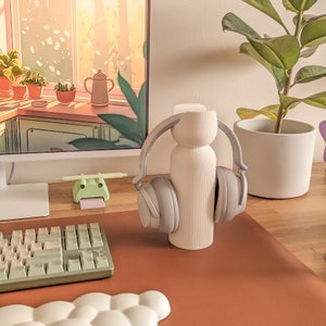 May include: A white ceramic headphone stand with a ribbed texture. The stand is holding a pair of grey over-ear headphones. The stand is on a brown wooden surface.