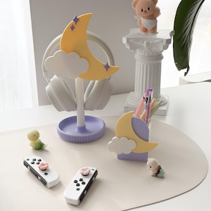 May include: A whimsical desk setup featuring a yellow crescent moon headphone stand with purple accents, white headphones, and a matching pen holder. Includes white game controllers with pink flower buttons, and small decorative figurines.