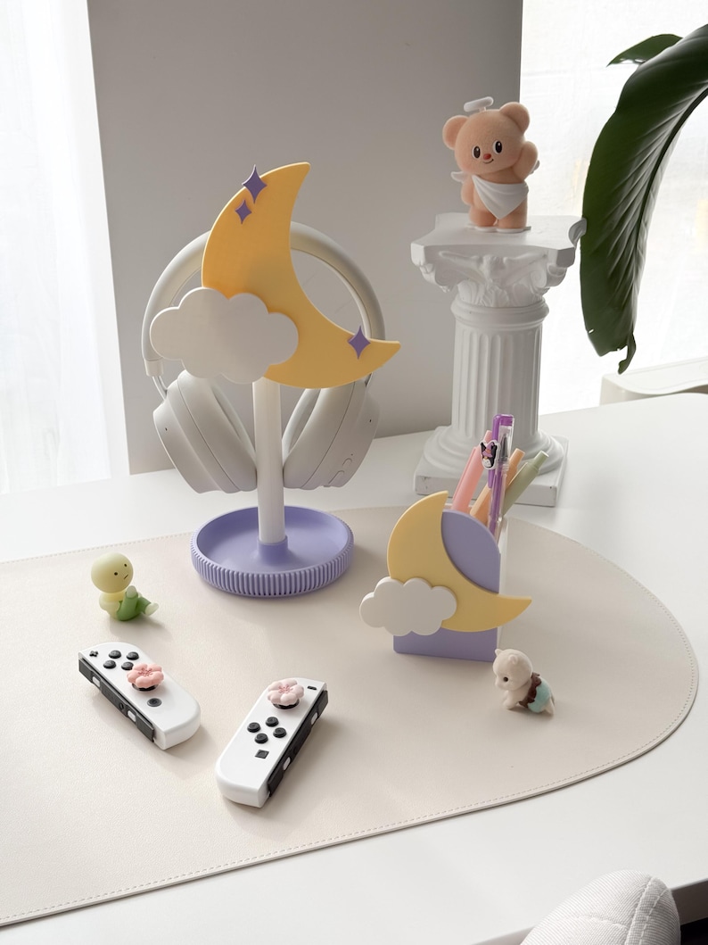 May include: A whimsical desk setup featuring a yellow crescent moon headphone stand with purple accents, white headphones, and a matching pen holder. Includes white game controllers with pink flower buttons, and small decorative figurines.
