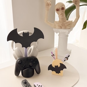 May include: A Halloween-themed desk organizer set featuring a black bat-shaped headphone stand, a black game controller holder, and a pen holder with a bat design. A skeleton statue is also present. The set includes gray and pink game controllers.