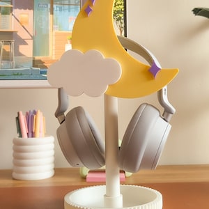 May include: A white headphone stand with a yellow crescent moon and a white cloud. The moon has purple stars on it. Two gray headphones are on the stand.