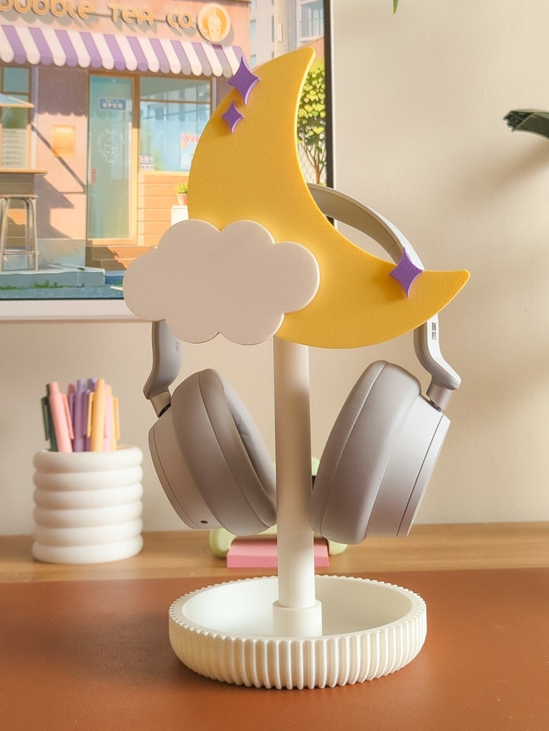 May include: A white headphone stand with a yellow crescent moon and a white cloud. The moon has purple stars on it. Two gray headphones are on the stand.