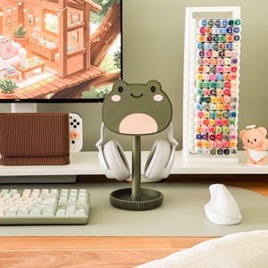 May include: A desk setup featuring a frog-shaped headphone stand holding white headphones. A white keyboard, mouse, and a marker holder filled with colorful markers are also visible. The desk has a green surface, and a computer monitor displays a cozy treehouse scene.