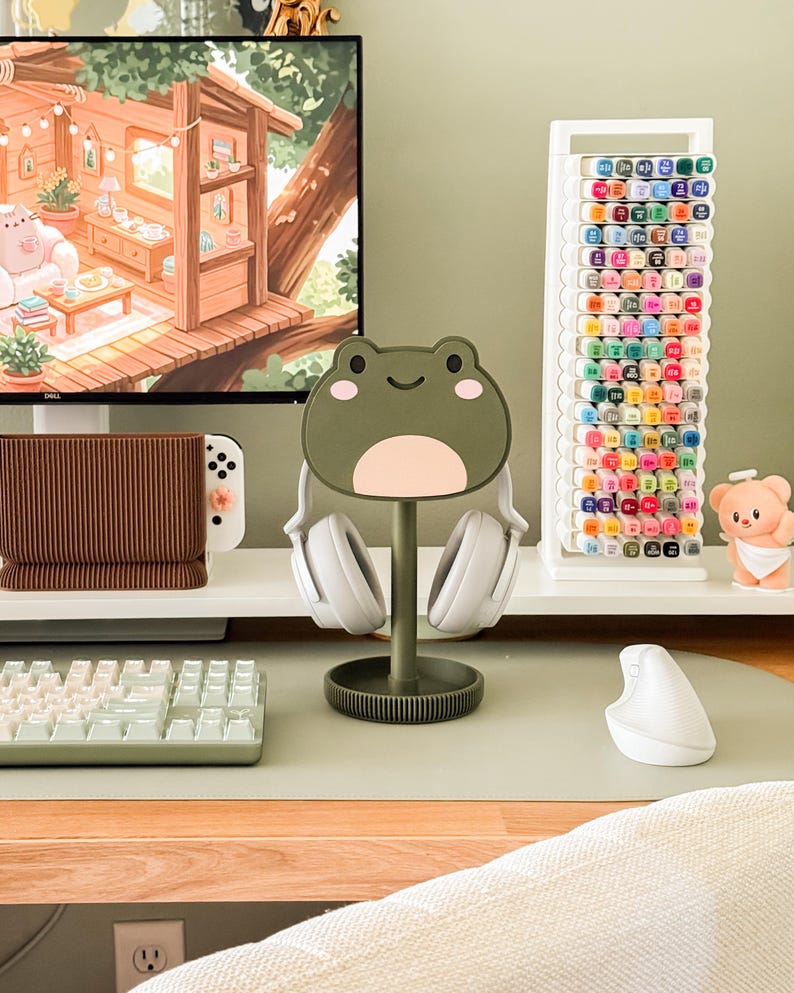 May include: A desk setup featuring a frog-shaped headphone stand holding white headphones. A white keyboard, mouse, and a marker holder filled with colorful markers are also visible. The desk has a green surface, and a computer monitor displays a cozy treehouse scene.