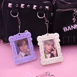 May include: Three decorative photo keychains in pastel colors: lavender, white, and pink. Each keychain features a small framed photo and a silver chain. The background includes a black bag with silver studs.
