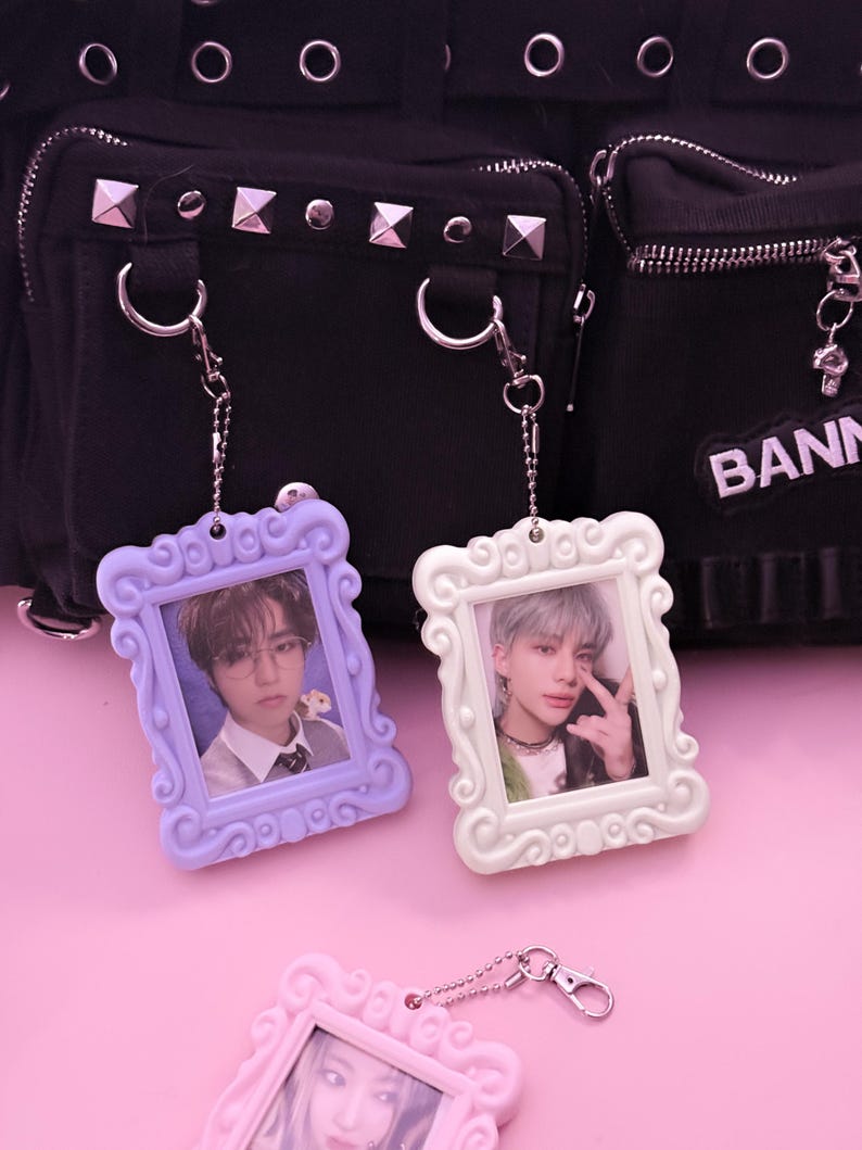May include: Three decorative photo keychains in pastel colors: lavender, white, and pink. Each keychain features a small framed photo and a silver chain. The background includes a black bag with silver studs.