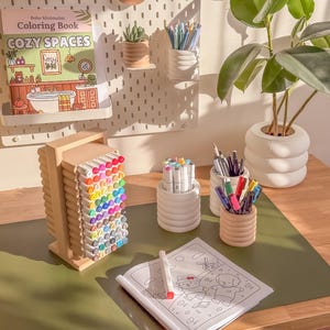 May include: A desk setup featuring a coloring book, various colored markers, and pens in decorative holders. A small succulent and a plant in a white pot are also visible. The coloring book is titled "Cozy Spaces."