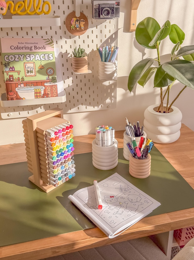 May include: A desk setup featuring a coloring book, various colored markers, and pens in decorative holders. A small succulent and a plant in a white pot are also visible. The coloring book is titled "Cozy Spaces."