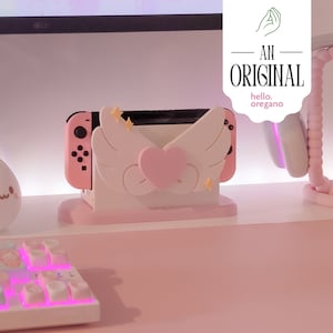Könnte beinhalten: Rosa Nintendo Switch Konsolenständer mit einem weißen und rosa Flügeldesign und einem rosa Herzen. Der Ständer hat eine kleine, weiße Basis.