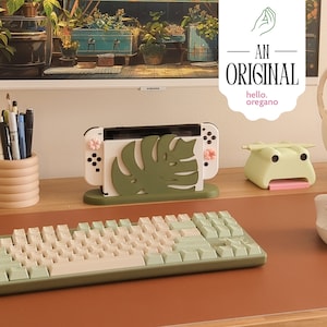 Peut inclure: Une console Nintendo Switch blanche avec un motif de feuille de monstera verte sur un support. La console est sur un bureau brun avec un clavier vert et blanc et un objet en forme de grenouille verte.