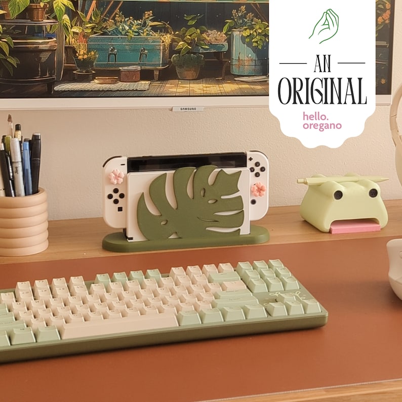 Peut inclure: Une console Nintendo Switch blanche avec un motif de feuille de monstera verte sur un support. La console est sur un bureau brun avec un clavier vert et blanc et un objet en forme de grenouille verte.