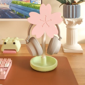 May include: A pink flower-shaped headphone stand with two gray headphones hanging from it. The stand is on a brown surface with a keyboard and a green frog figurine.