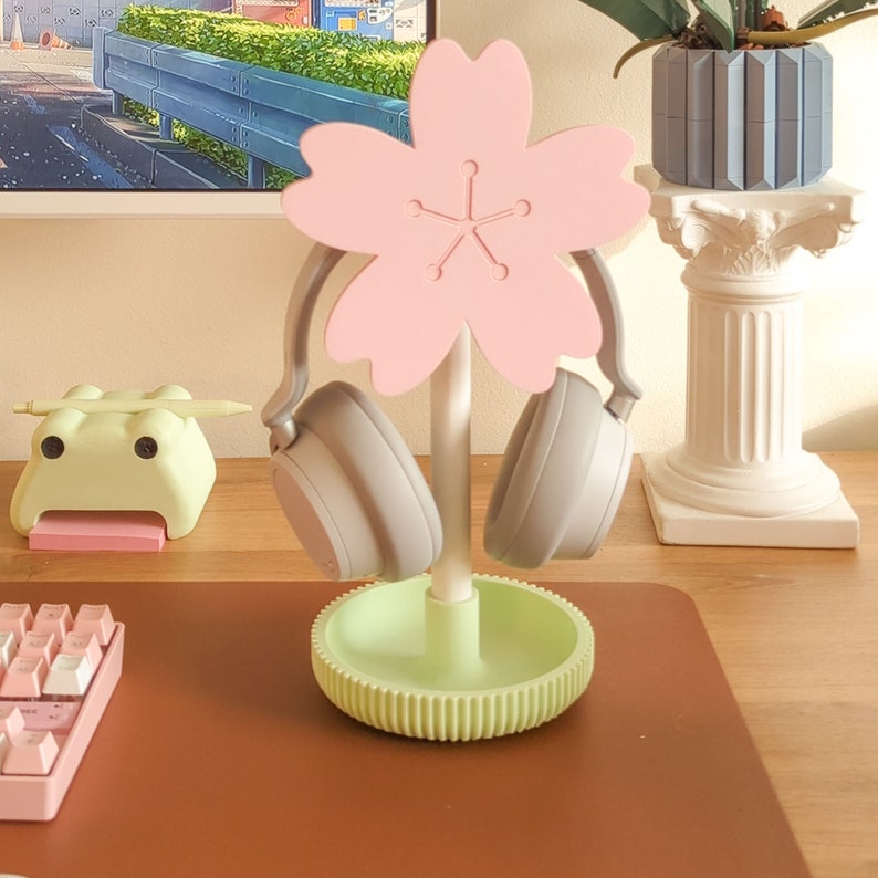 May include: A pink flower-shaped headphone stand with two gray headphones hanging from it. The stand is on a brown surface with a keyboard and a green frog figurine.